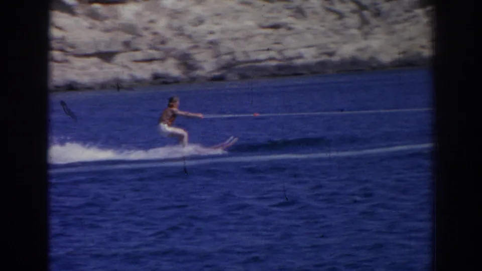 A male water skier in dark swim trunks rides on a single ski, with blue water and a rocky Izmir coastline behind him.