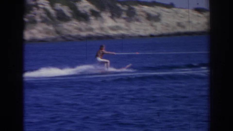 Final frame of a 1962 Izmir water-skiing shot, with a male skier towed across blue water against a rocky coast.