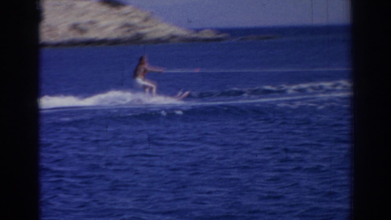 Water skier in white pants crosses a blue sea near a rocky coastline.