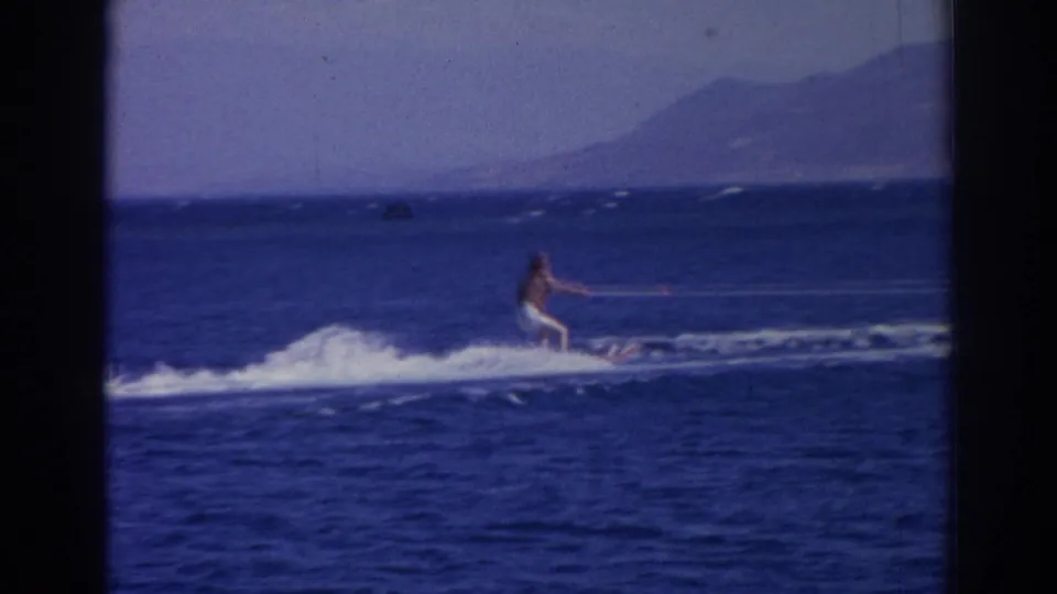 A water skier travels across the bright blue sea with distant mountains visible in 1962 Izmir home-movie footage.