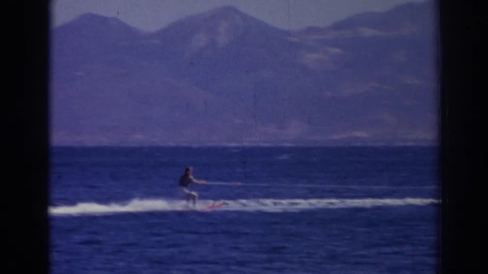 A distant skier in white pants cuts across the blue sea with hazy mountains behind in this 1962 Izmir footage.