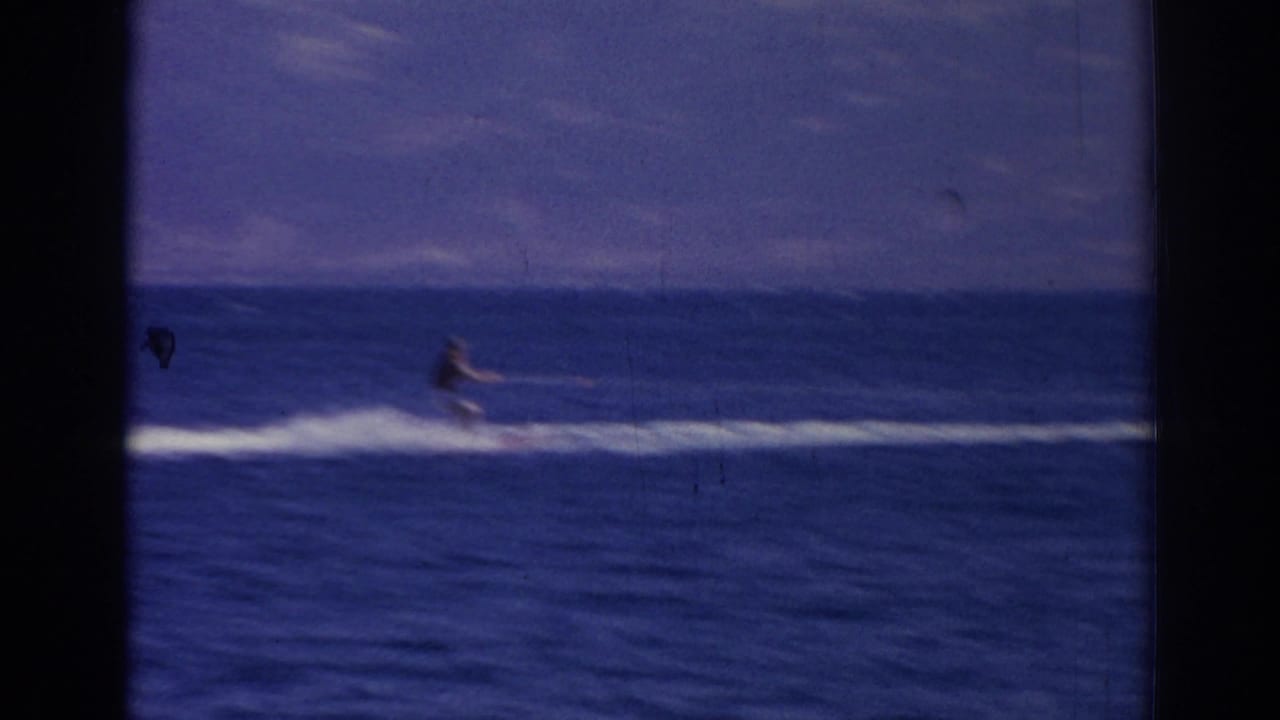Person riding a small watercraft across blue water with a white wake and distant mountains.