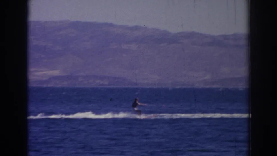 1962 Izmir home-movie frame showing a distant rider low on the water, with a pale wake and layered mountains behind.