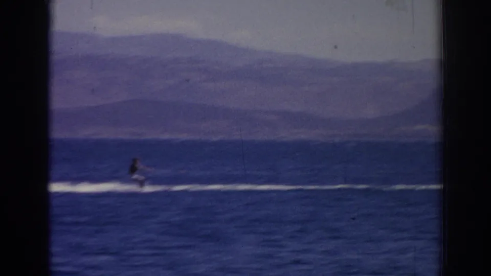 Final 1962 Izmir frame of the rider continuing across dark blue water, with white spray and a hazy mountain backdrop.