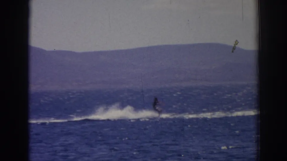 In 1962 Izmir-area home-movie footage, a distant water skier moves across sunlit blue water below a hazy mountain ridge.