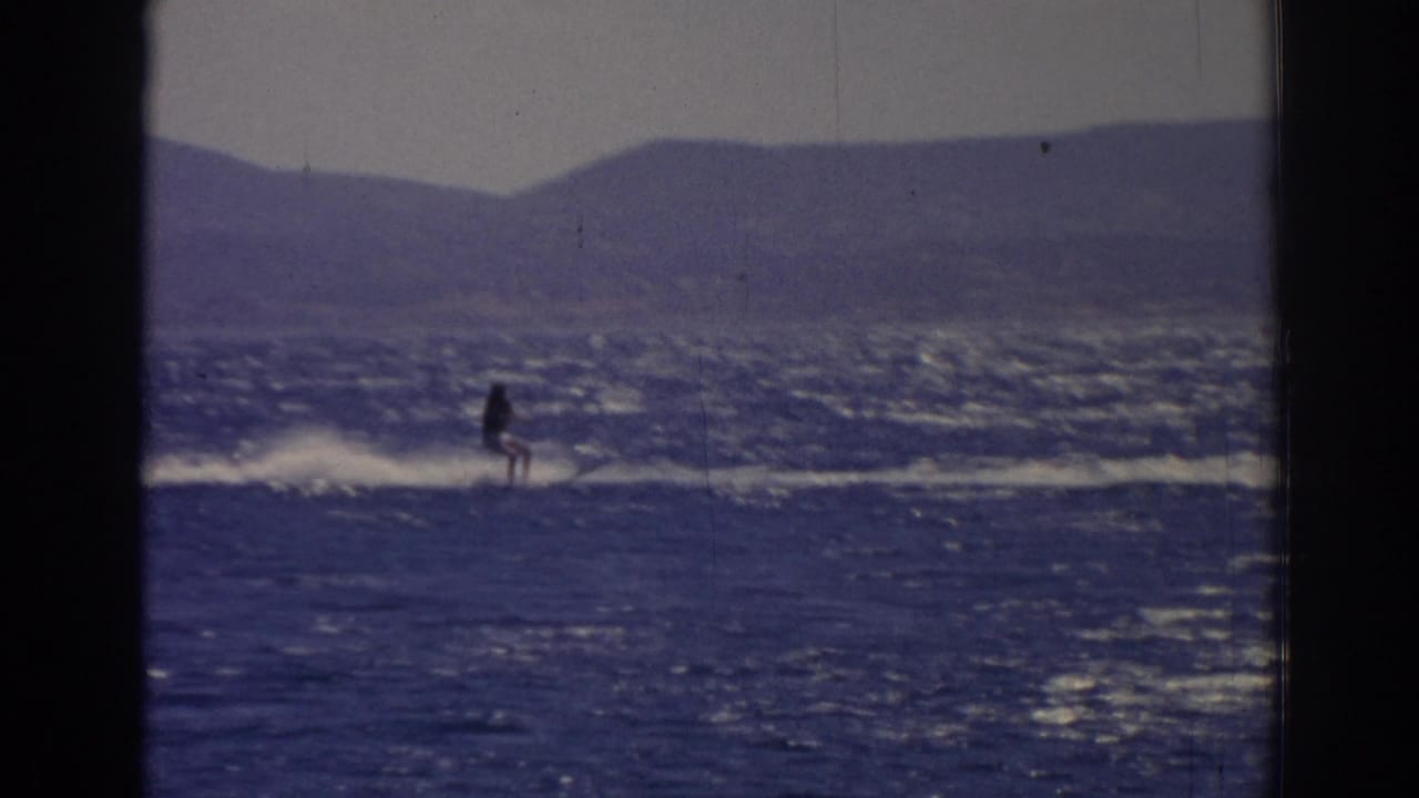 A distant skier continues across the choppy lake in Izmir, Turkey, with sun glare on the water and mountains behind.