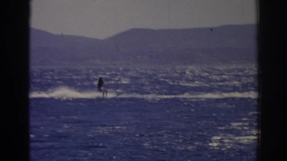 A distant skier continues across the choppy lake in Izmir, Turkey, with sun glare on the water and mountains behind.