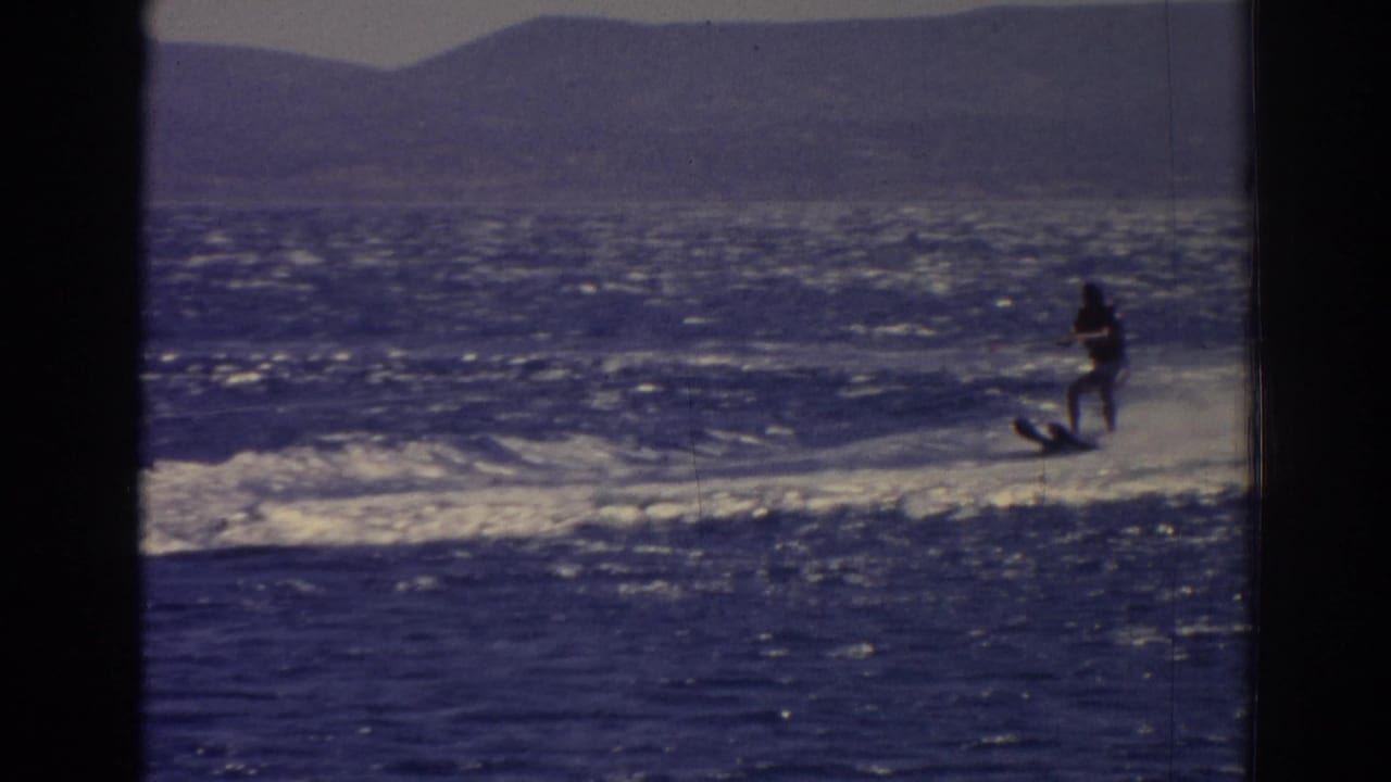 A distant skier in dark clothing and light shorts rides over a white wake on blue water in the 1962 Izmir footage.