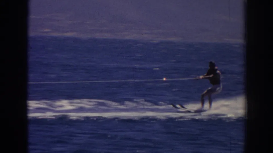 A distant skier leans back on water skis while a taut tow rope stretches across the frame over blue water.