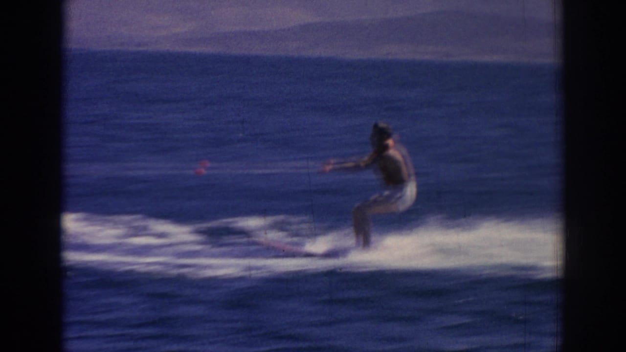 A distant adult waterskier in a dark top and light shorts rides across blue water near Izmir in the 1962 home movie.