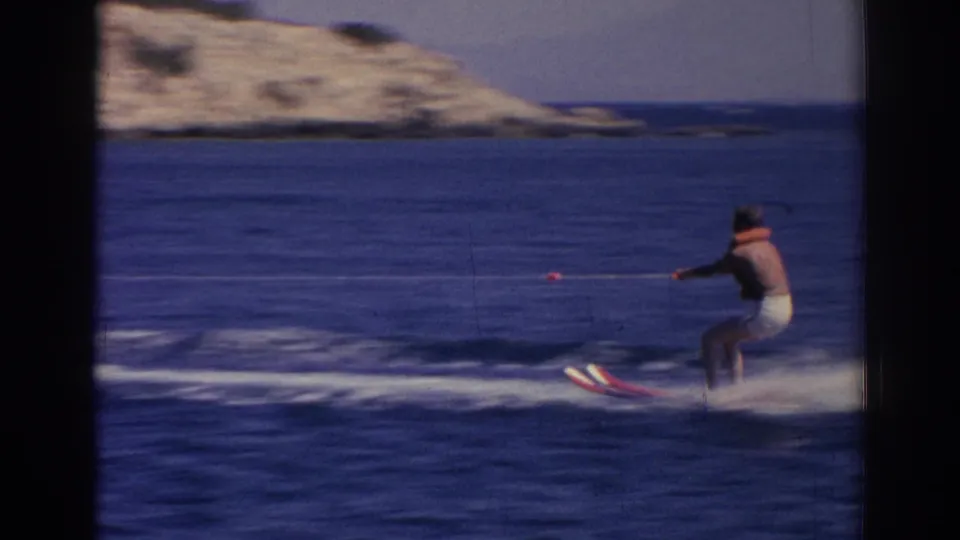A man on water skis is pulled across open blue water near a pale shoreline in the supported 1962 Izmir home-movie footage.