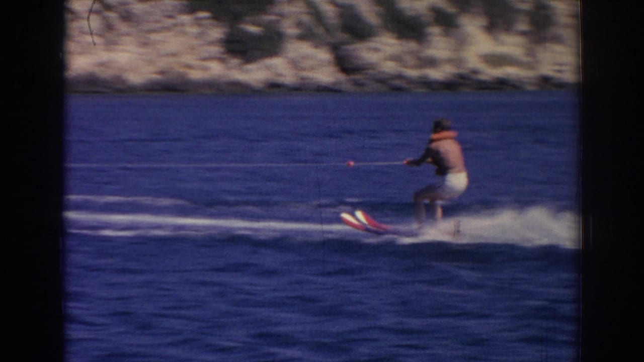 Man water skiing behind a motorboat on blue water near a rocky shoreline.