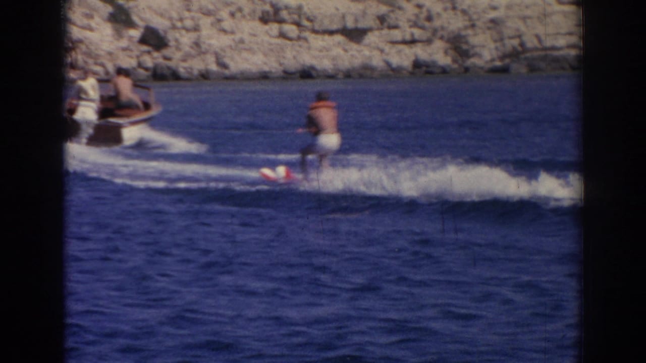 1962 Izmir, Turkey frame with a small motorboat towing a water skier across the sea near a rocky coast.