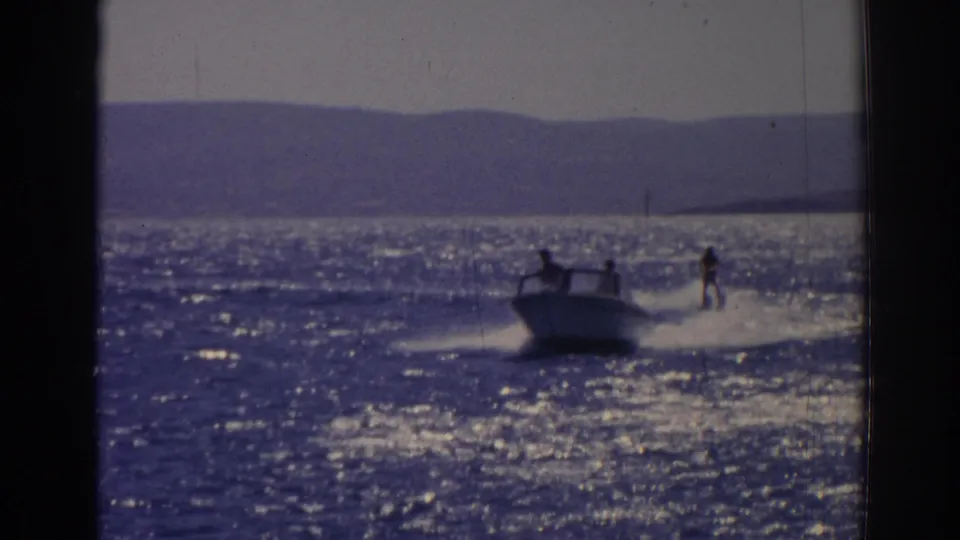 1962 Izmir, Turkey frame showing a water skier behind a motorboat on reflective blue water with hills in the distance.