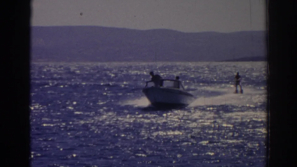 1962 Izmir home-movie frame showing a motorboat towing a water skier across sunlit coastal water with hills in the background.