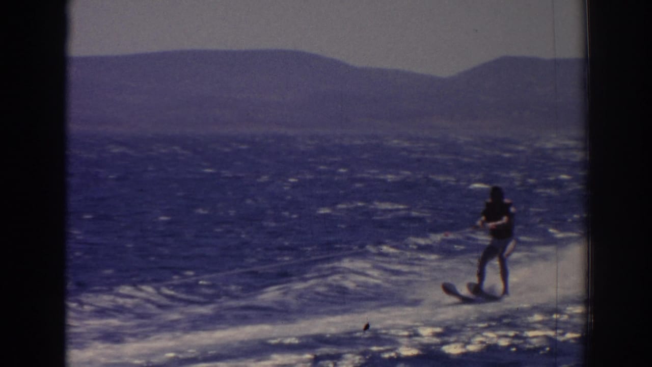 Water skier gliding over a boat wake on blue water near distant hills.