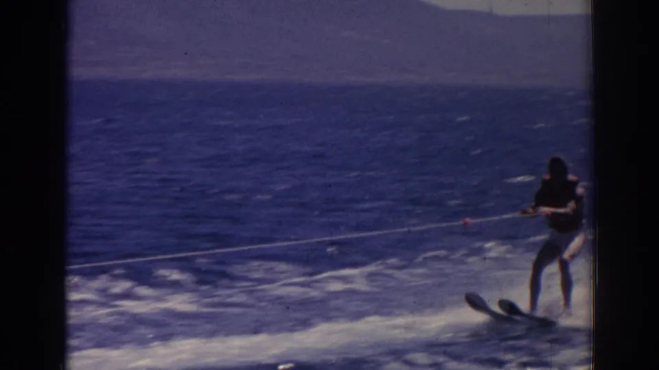 A closer 8mm frame shows the skier leaning slightly while holding the tow rope, with open blue water and wake surrounding him.