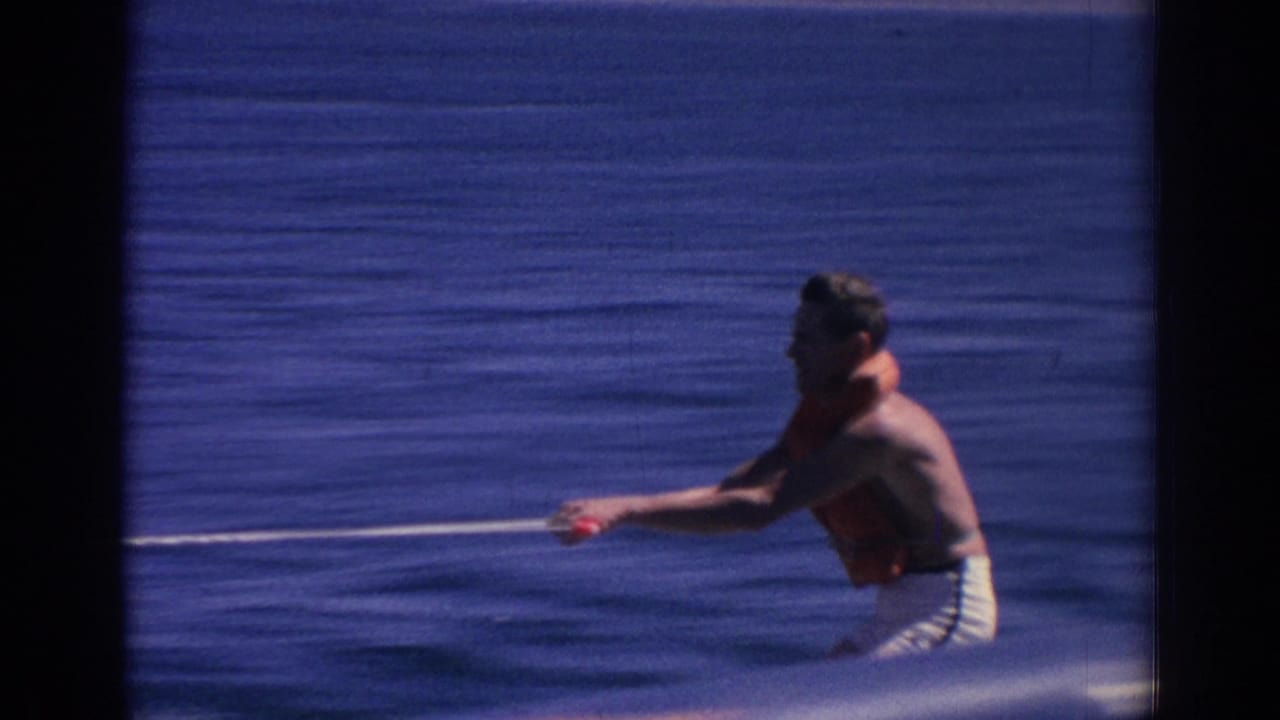 Man in orange flotation gear holds a tow rope while water skiing on blue water.