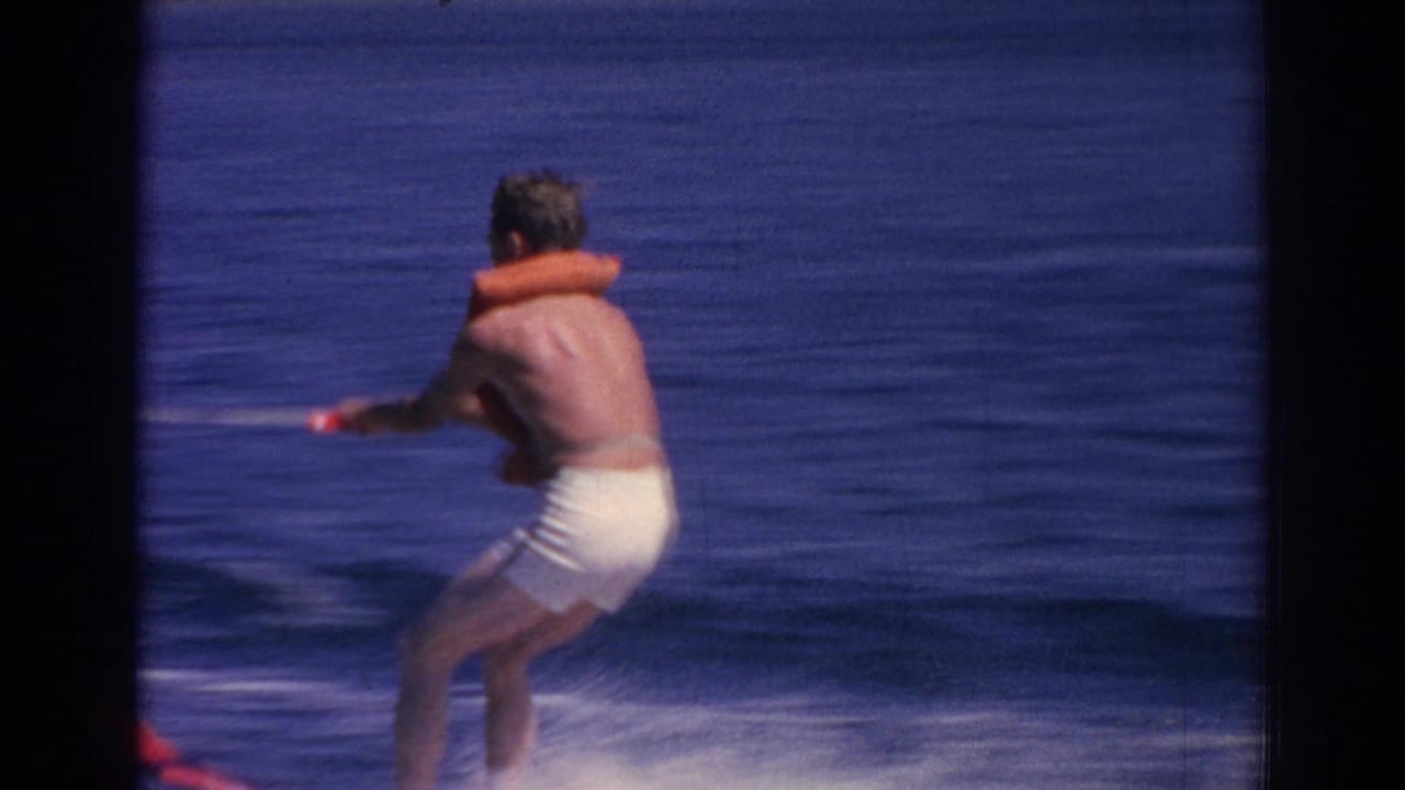 A man water skis across blue water near Izmir, wearing white swim trunks and orange flotation gear as he follows the tow line.