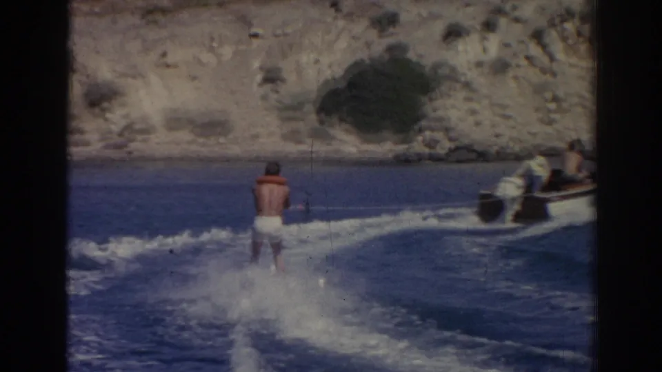 From a wider view, the water skier follows the motorboat across blue water with rocky cliffs in the background near Izmir.