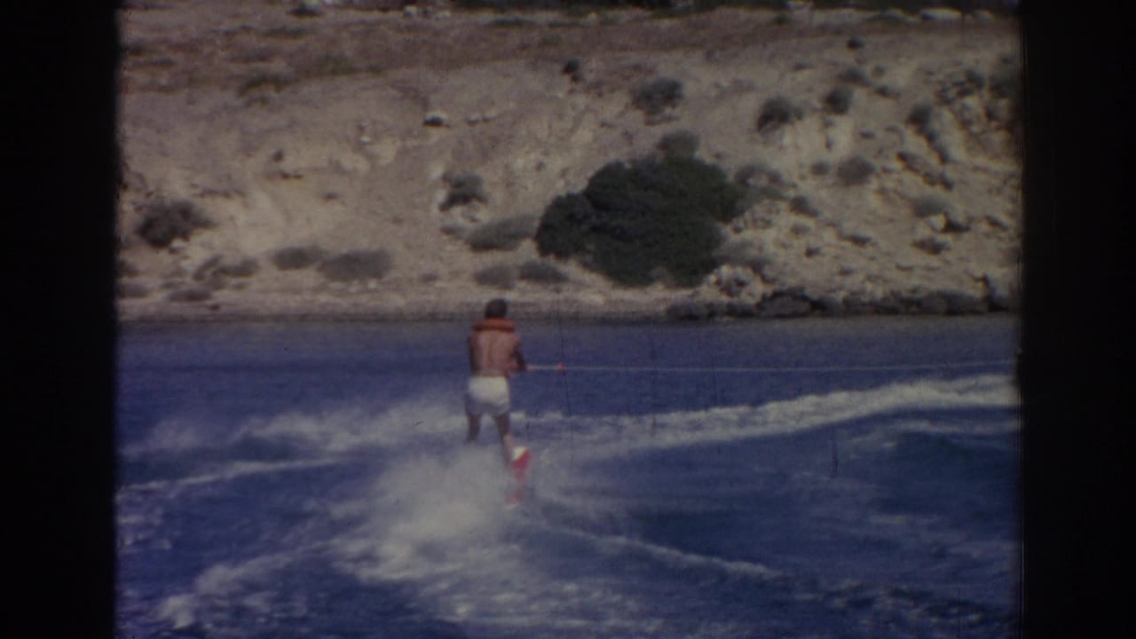 In 1962 Izmir, Turkey footage, a man in swim trunks and a life vest rides behind a boat across blue water near a rocky shore.