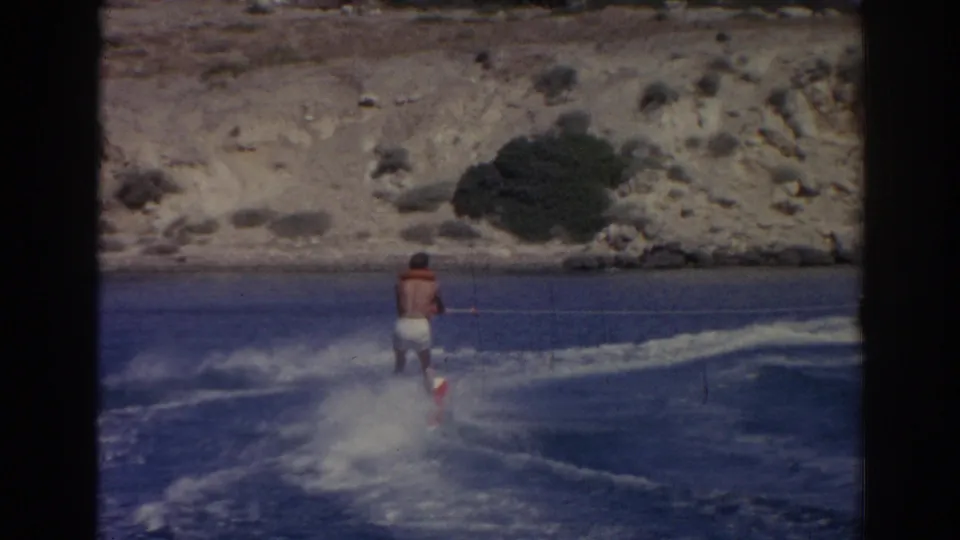 In 1962 Izmir, Turkey footage, a man in swim trunks and a life vest rides behind a boat across blue water near a rocky shore.