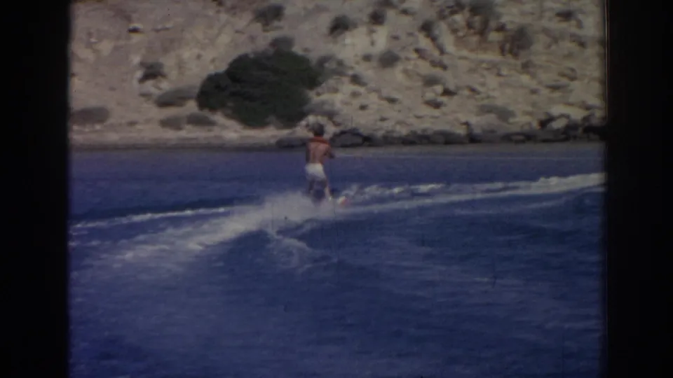 A man rides over the deep blue sea behind a boat, with the arid Izmir coastline visible in the background.