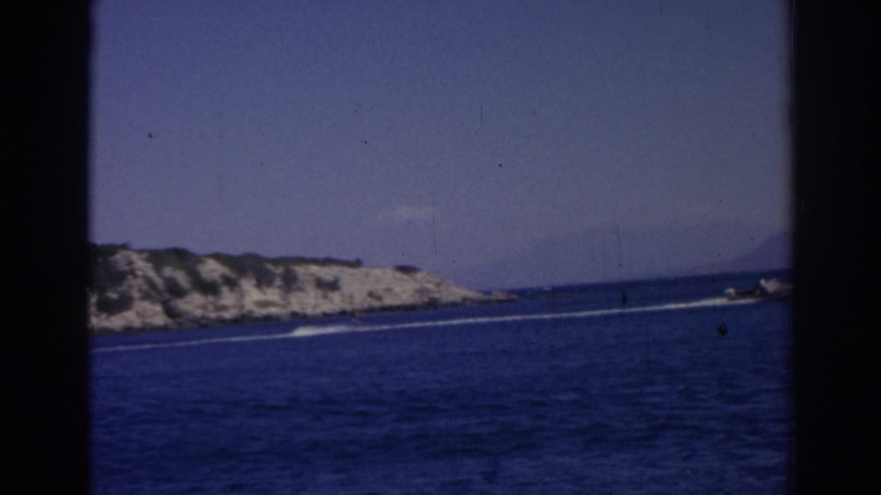 Small motorboat wake crossing deep blue sea near a rocky coastline under a pale sky.