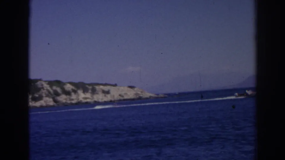 1962 Izmir, Turkey home-movie frame showing blue sea, a distant motorboat wake, rocky shore, and pale sky.