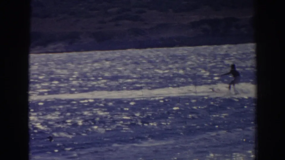 Home-movie frame from Izmir in 1962 showing a distant water skier tracking along the shore across bright water.