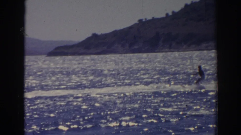 1962 Izmir footage frame with a distant skier at lower right, a white wake, sparkling sea, and hilly shore.