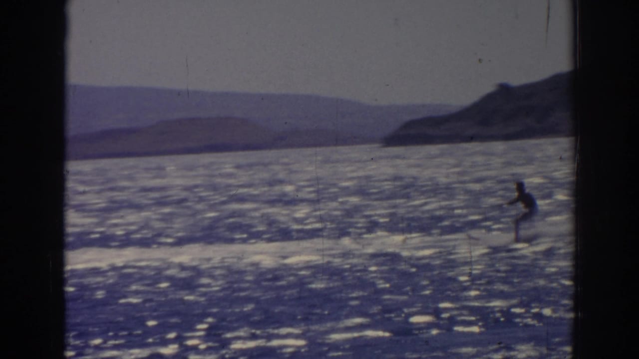 Distant water skier crossing bright open water with hills in the background.