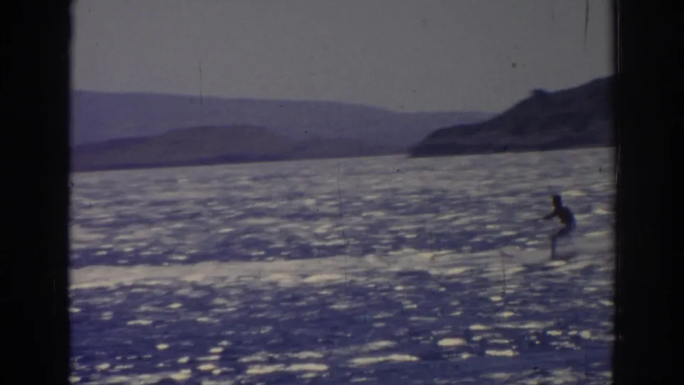 1962 Izmir home-movie frame showing a distant water skier on shimmering open water, seen through dark film or window edges.