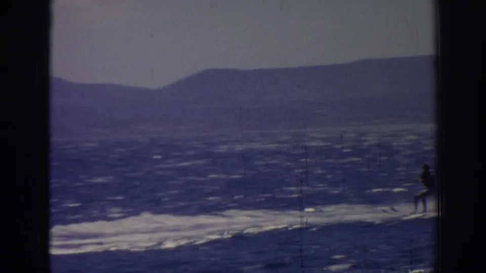 A distant skier appears at the frame edge in this Izmir 8mm home-movie view, with hills and rippled water behind.