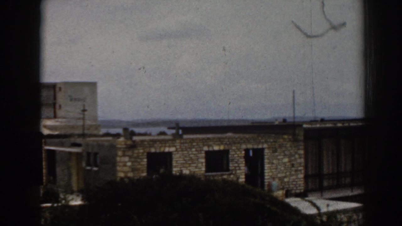 Distant stone apartment block seen through a dark window edge with sea and cloudy sky behind it.