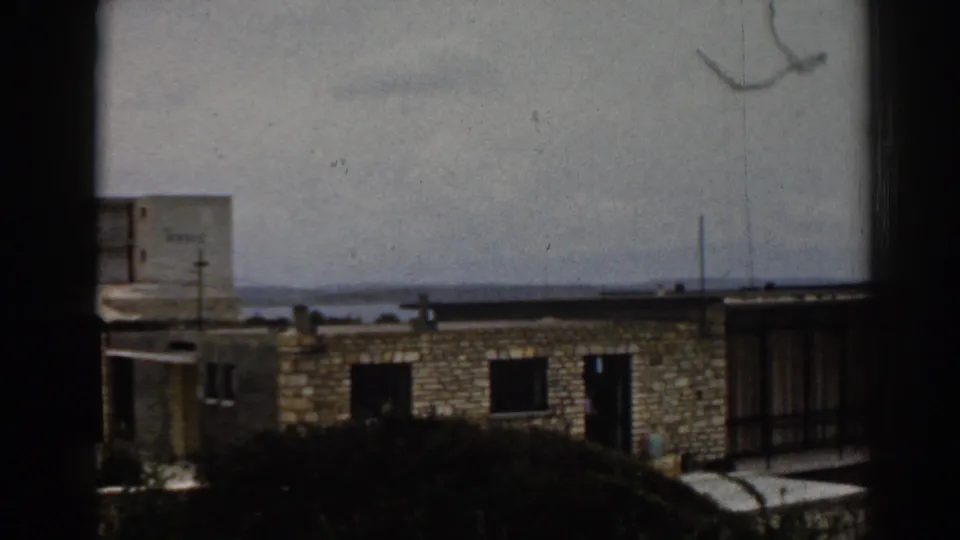 1962 Izmir view of a stone apartment block framed by a dark window edge, with a flat roofline, shrubs, sea horizon, and overcast sky.