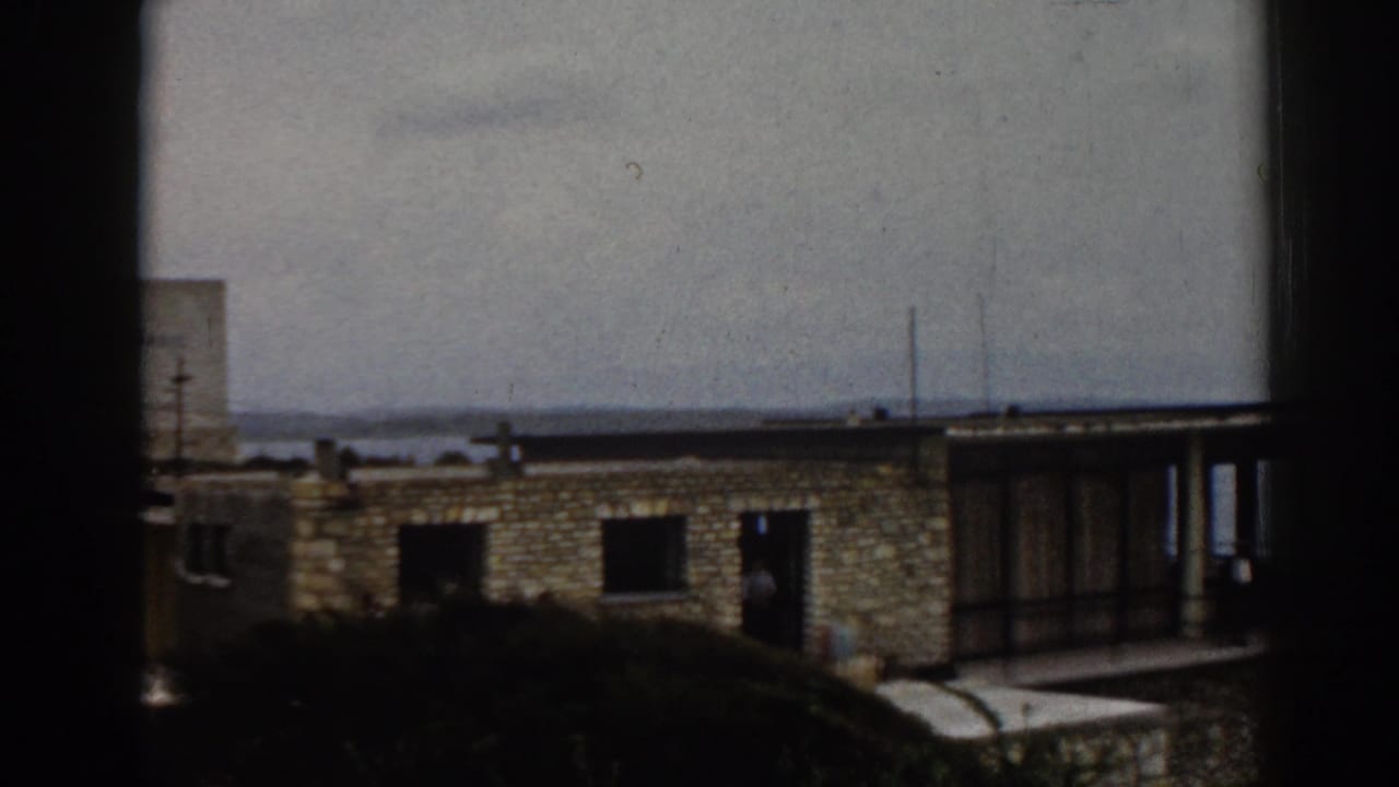 Stone building and glazed balcony area viewed across shrubs, with a coastal horizon in the background.