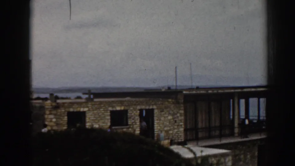 The camera holds on a distant 1962 Izmir apartment block, with dark window framing, stonework, open windows, vegetation, and the sea behind.