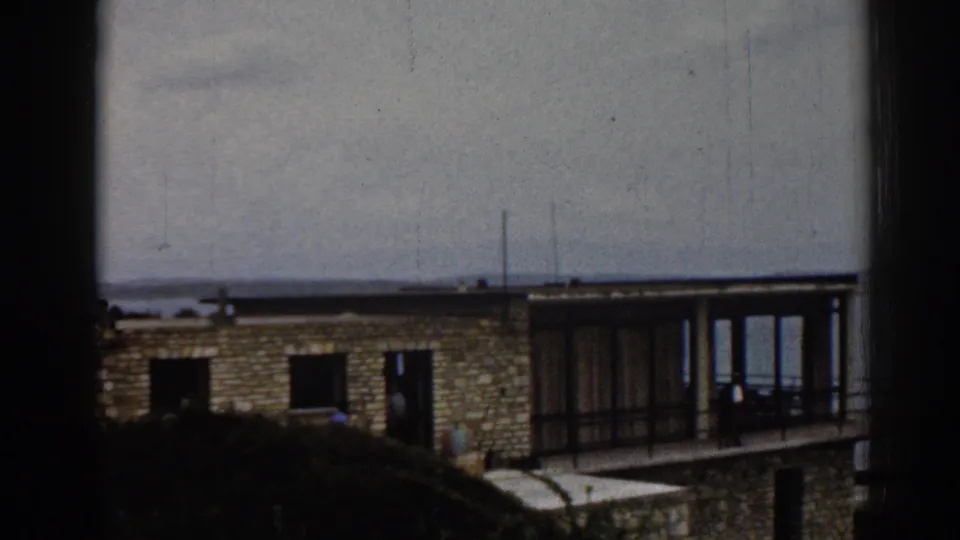 A distant 1962 Izmir view shows the stone apartment block, glazed balcony section, shrubs, sea horizon, and tiny people near the building.