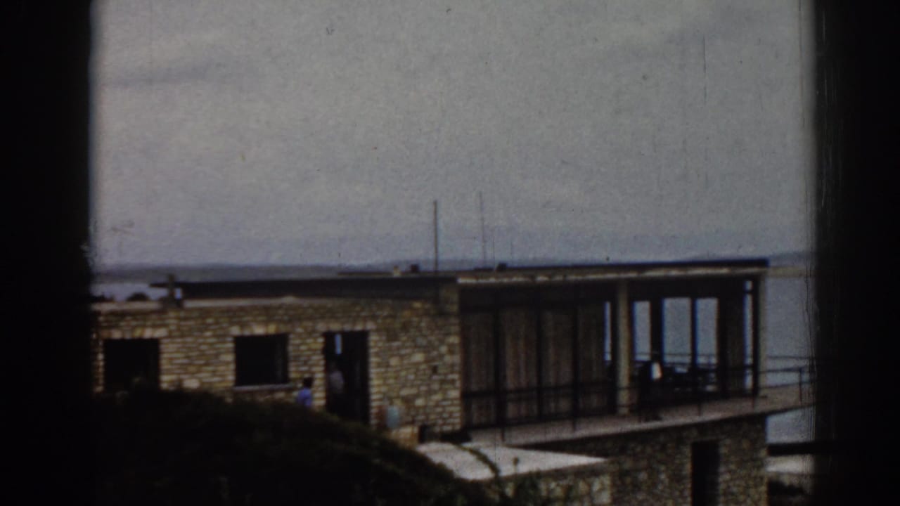Stone house with glass balcony beside a calm lake, partly obscured by dark frame edges.