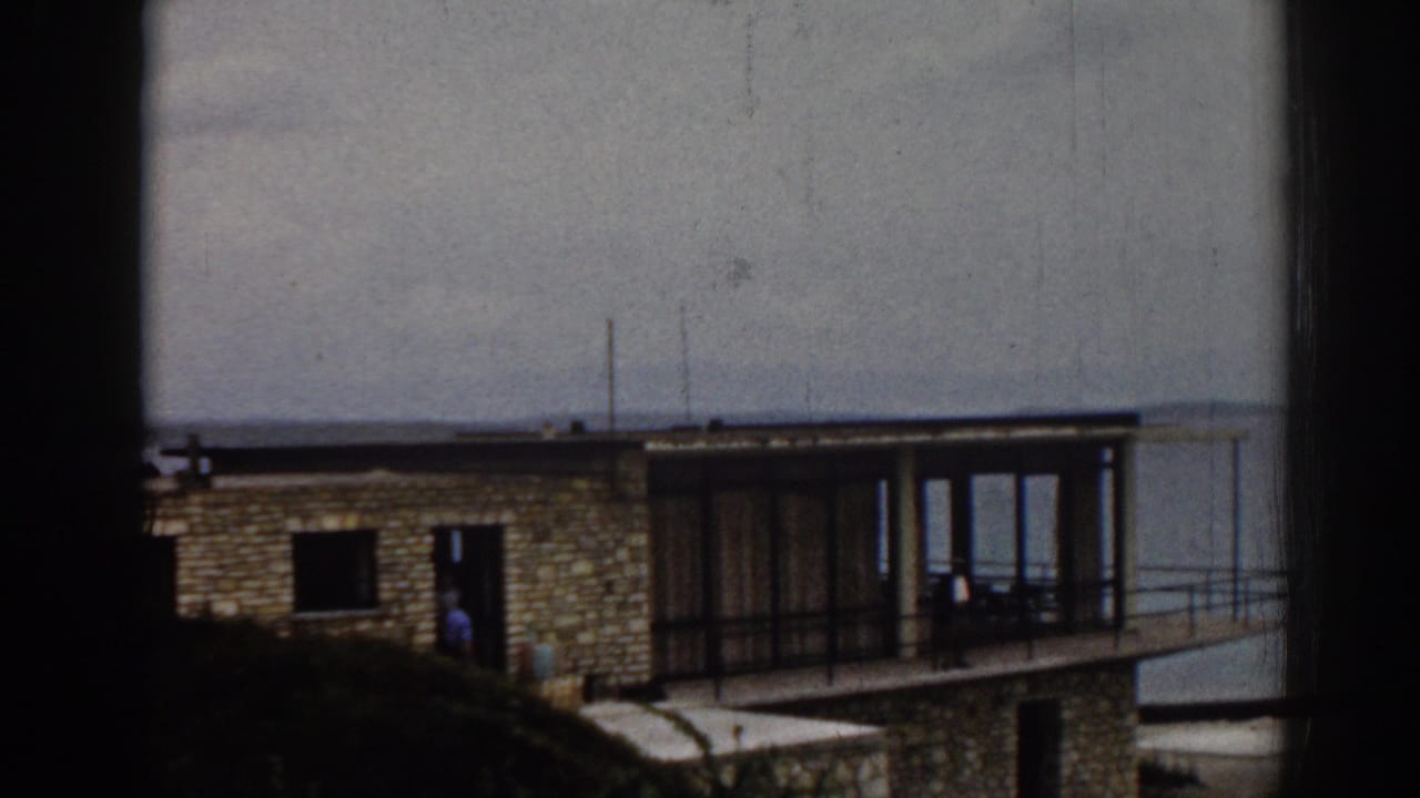 1962 Izmir frame of a stone house by the lake, with a small figure near the entrance and a glass-walled balcony facing the water.