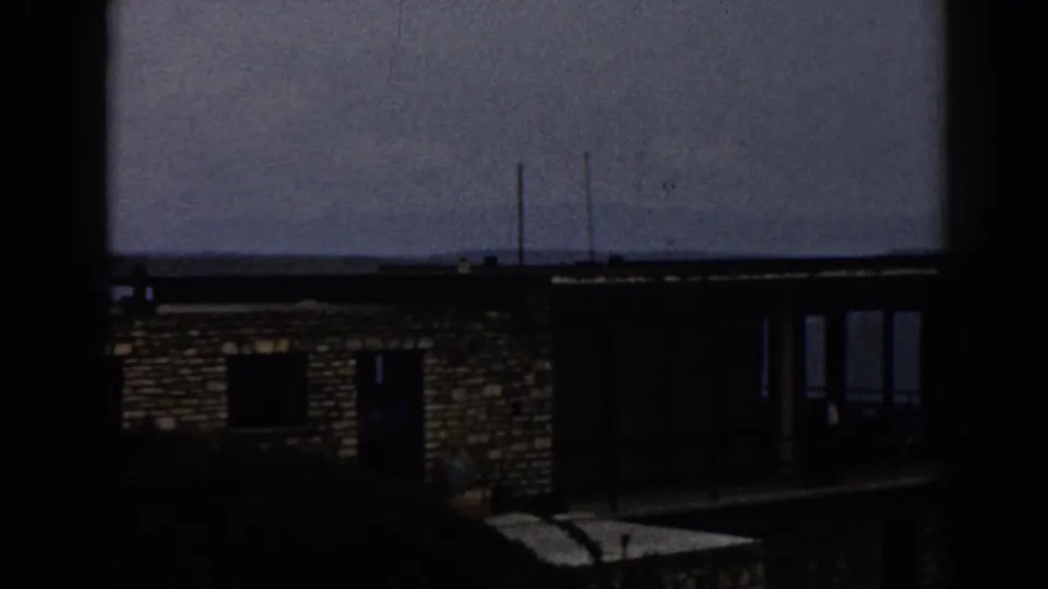 Underexposed 1962 Izmir home-movie frame showing the lakeside stone house, dark windows, balcony structure, and water in the background.