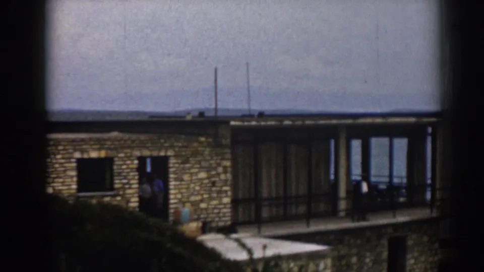 1962 Izmir frame showing a stone lakeside house, a person at the entrance, terrace railings, and a broad hazy lake view.
