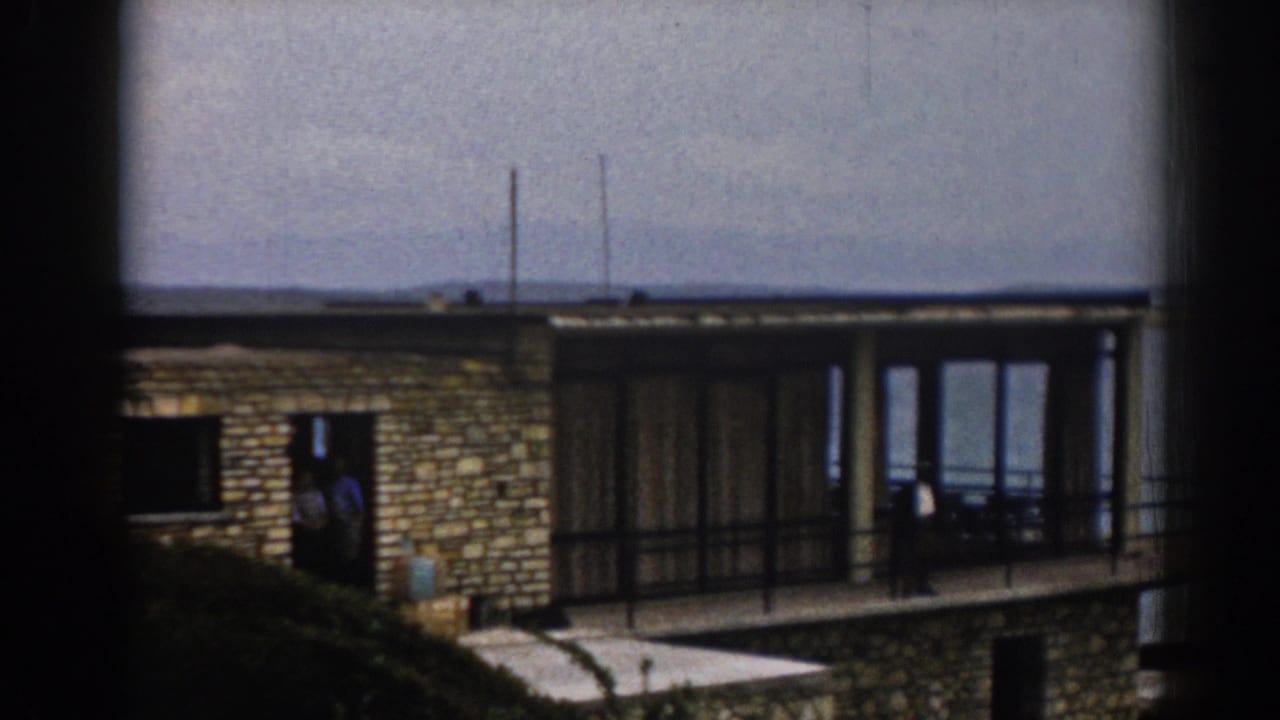 Seaside house with stone facade, balcony, glass doors, and two people near the entrance and railing.