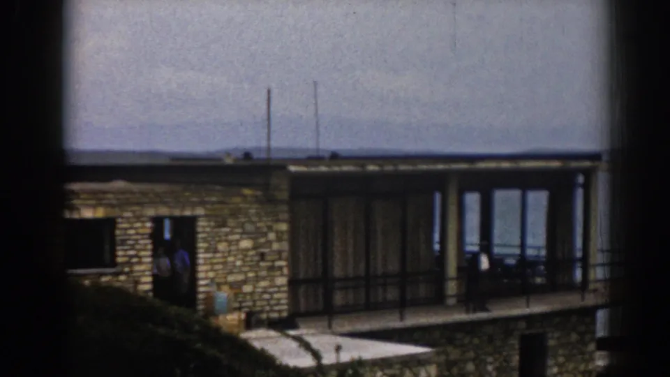 1962 Izmir home-movie frame showing a modern seaside house with stone facade, balcony railings, glass doors, and two distant figures.