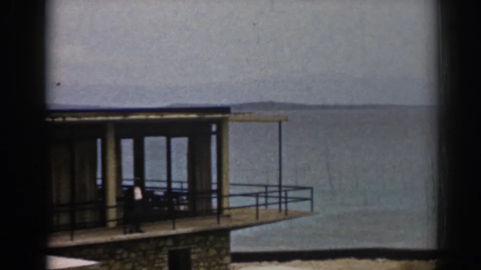 1962 Izmir home-movie view of a glass-fronted balcony on a coastal house, with calm water and faint hills in the background.