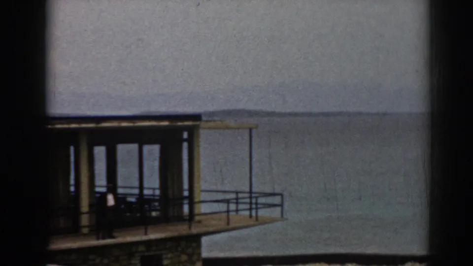 1962 Izmir frame showing the house balcony and glass doors from an oblique angle, with calm sea and hazy shoreline beyond.