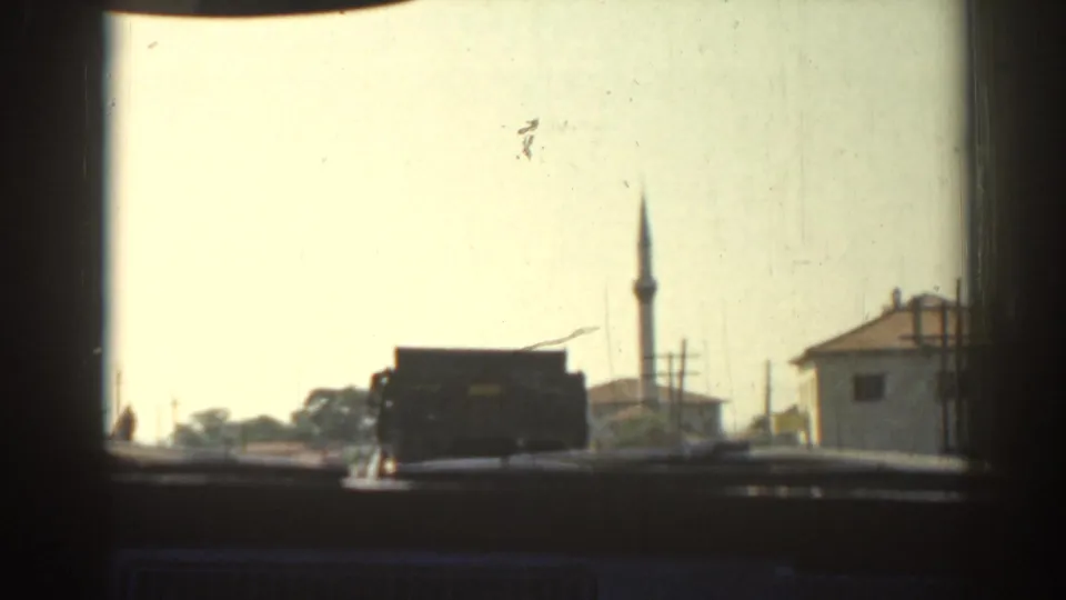 1962 Izmir street view filmed through a vehicle window, with a mosque minaret, low buildings, poles, and hazy sky.