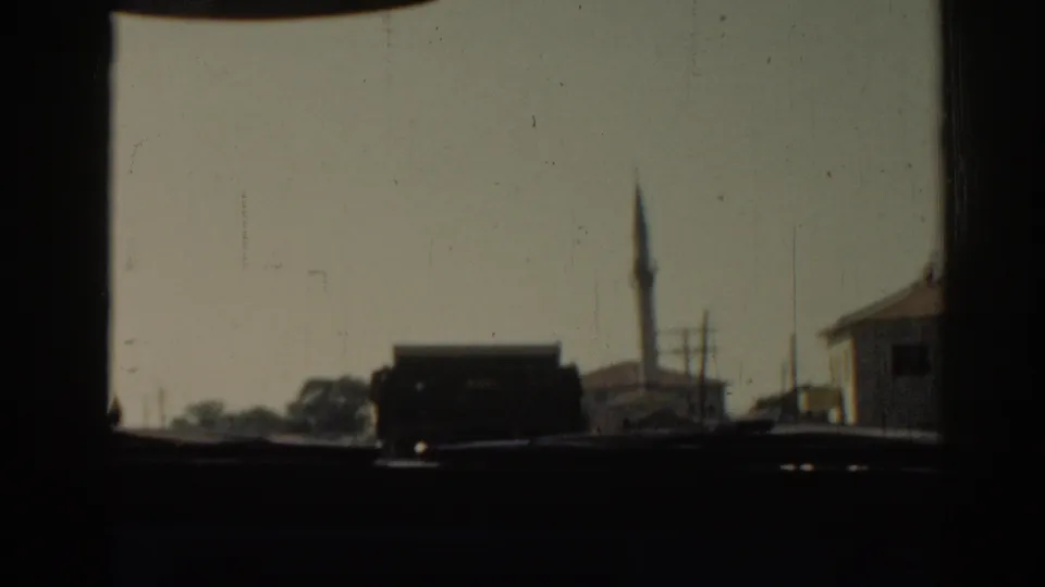 1962 Izmir home-movie frame from inside a moving vehicle, showing a minaret, roadside buildings, and hazy light.