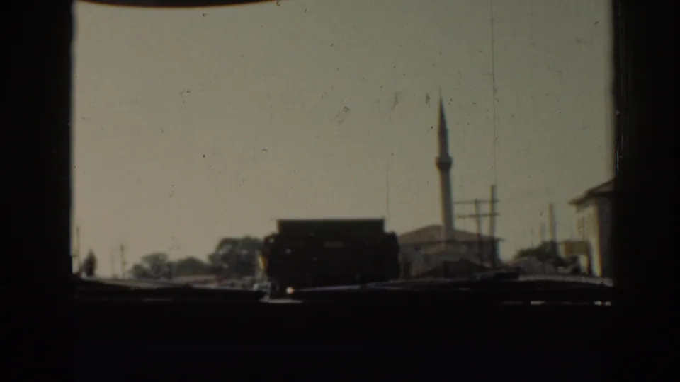 1962 Izmir car-window view showing a dark truck, low buildings, utility poles, and a mosque minaret in soft 8mm color.